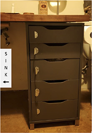 Stack of five drawers with thin aluminum handles rotated ninety degrees counterclockwise and sticking straight out from the drawer face. The top and bottom drawers have had the handles broken off. A label indicates the sink is located to the left of the drawers.